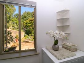 A small indoor room with a window overlooking a garden a white counter with a potted flower a basket holding toilet paper and a wall-mounted corner shelf at Garden Cottage in Black Bourton near Burford