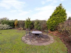 An outdoor seating area with a round mosaic table and four metal chairs on a stone patio surrounded by grass and shrubs at The West Wing in Bodfari