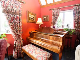A room with a wooden piano and a matching bench with patterned cushions next to windows with patterned curtains and framed paintings on the wall at The West Wing in Bodfari