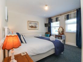 A bedroom with a bed, bedside table with lamp, chair, dressing table with mirror, windows with blinds and curtains, and wall art at Sea View apartment in Borth-Y-Gest