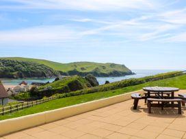An outdoor patio with a round wooden table and benches overlooking green hills water and houses at 2 Sea Gardens in Hope Cove