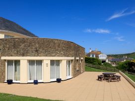 A stone and stucco house with large windows and a patio with a round wooden table and benches at 2 Sea Gardens in Hope Cove