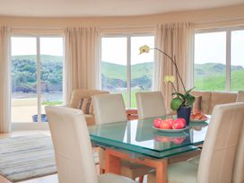 A dining area with a glass table and beige chairs with a flower pot and fruit plates near large windows showing green hills at 2 Sea Gardens Hope Cove