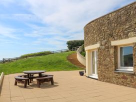 An outdoor patio area with a round wooden picnic table and curved stone building with steps leading up a grassy hill at 2 Sea Gardens in Hope Cove