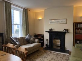A living room with a beige sofa with cushions near a window with curtains a black fireplace with two elephant figurines on the mantle a wooden bookshelf filled with books and a wooden dining table with chairs at Sant Seiriol in Beaumaris