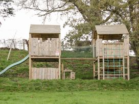 A wooden playground structure with two towers connected by a rope bridge and a slide on the left at Cymryd Uchaf in Conwy