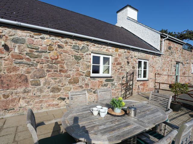 An outdoor patio with a wooden table and chairs with cups and a teapot against a stone cottage wall at Tan Twr - Chellow Cottage in Dwyran