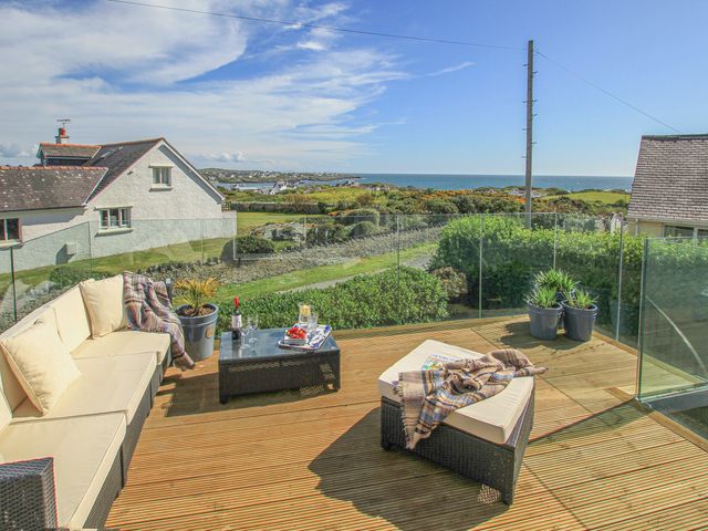 A wooden deck with outdoor seating and a glass table overlooking greenery and the sea at Curragho in Trearddur Bay
