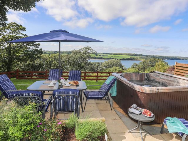 An outdoor patio with a dining table and chairs under a blue umbrella next to a hot tub and a small side table with fruit overlooking water and greenery at Gwylan Uchaf in Y Felinheli