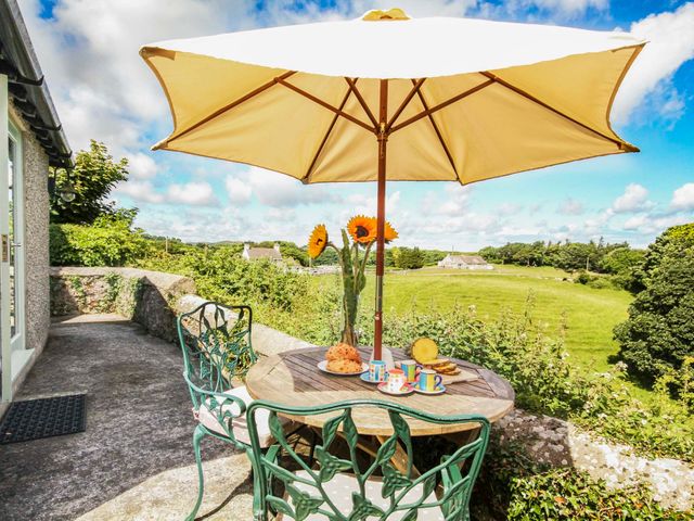 An outdoor patio with a round wooden table and green metal chairs under a yellow umbrella overlooking green fields at Maes y Cregyn in Marianglas