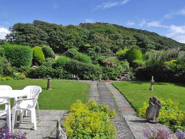 A garden with green bushes stone statues and a white table with plastic chairs at Mor Edrin in Criccieth