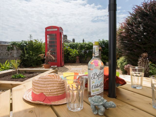 A wooden picnic table with a straw hat bottle and glasses in a garden with a red phone booth and plants at Swn y Mor Sound of the Sea in Criccieth