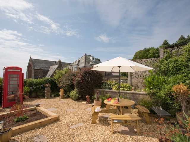 A garden with a round wooden picnic table and umbrella next to a red telephone box and plants at Swn y Mor Criccieth