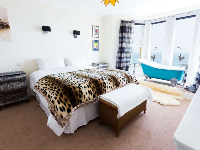 A bedroom with a bed a leopard print blanket bedside tables a blue bathtub and large windows at Bay View - Vista Marina in Criccieth