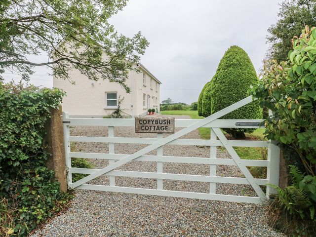 A white wooden gate with a sign reading Copybush Cottage leading to a gravel driveway and a cream house surrounded by trimmed bushes at Copybush Cottage in Lawrenny