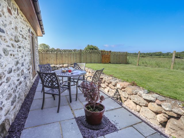 An outdoor patio with a round table and four chairs next to a stone wall and a wooden fence enclosing a grassy yard at Y Bwthyn in Brynsiencyn