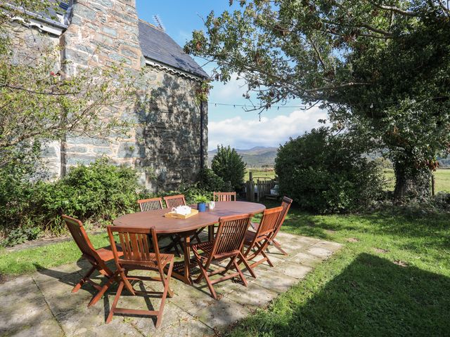 An outdoor wooden dining table with eight chairs on a stone patio next to a stone building with greenery and trees at Glyn Morfa in Harlech