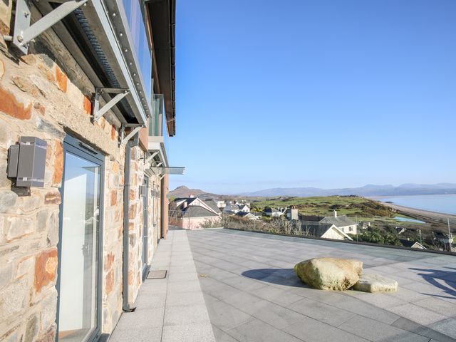A stone building with glass doors and a tiled terrace overlooking houses hills and a beach at Hafod Y Mor in Criccieth