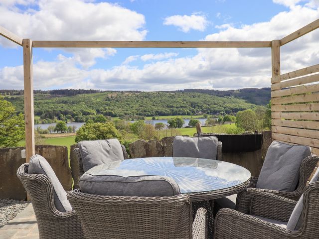 An outdoor seating area with wicker chairs and a glass table overlooking a lake and green hills at Two Lake View in Coniston