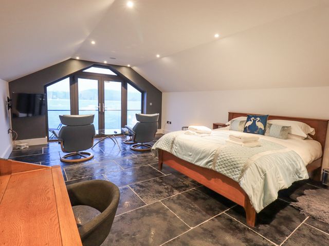 A bedroom with a bed two chairs a desk and large windows overlooking water at The Boat House at Louper Weir in Bowness-On-Windermere