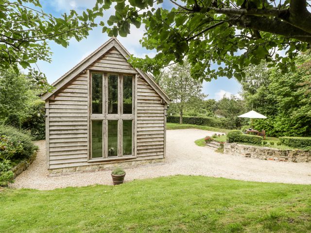 A wooden building with large vertical windows next to a gravel driveway and garden with an outdoor seating area and umbrella at The Barn at Frog Pond Farm Antsy near Tisbury