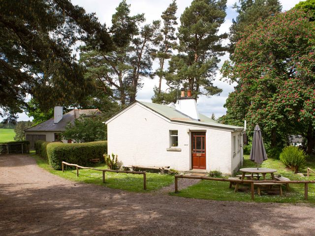 A small white house with a brown door surrounded by trees and green grass with a picnic table and umbrella at The Bungalow in Lhanbryde