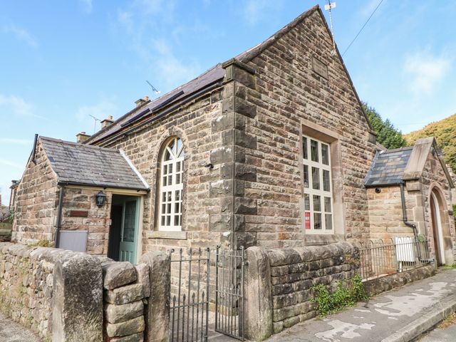 A stone building with arched windows and two small porches surrounded by a low stone wall and metal gates at Nine Ladies in Birchover