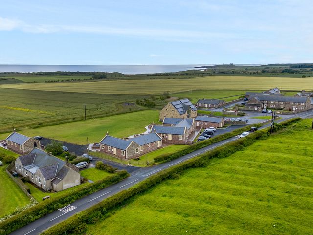 A cluster of stone buildings with parked cars along a road near green fields and the coast at The Retreat in Embleton