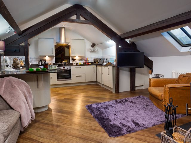 An open kitchen and living area with white cabinets wooden beams a brown leather chair and a purple rug at Hayloft in Windermere