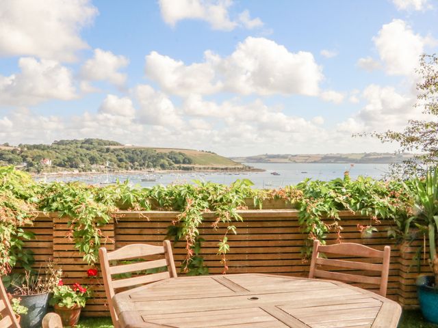An outdoor patio with wooden table and chairs overlooking a body of water with boats and green hills at Through The Porthole in Falmouth