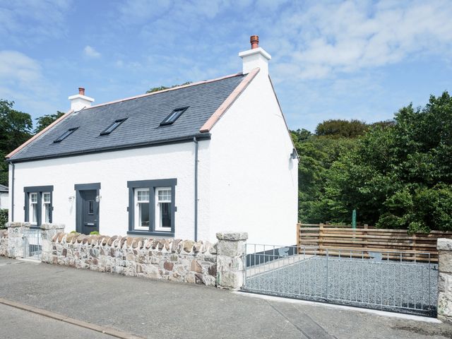 A white house with a dark roof and stone fence next to a gravel driveway and trees at Squirrel Cottage in Kirkcolm