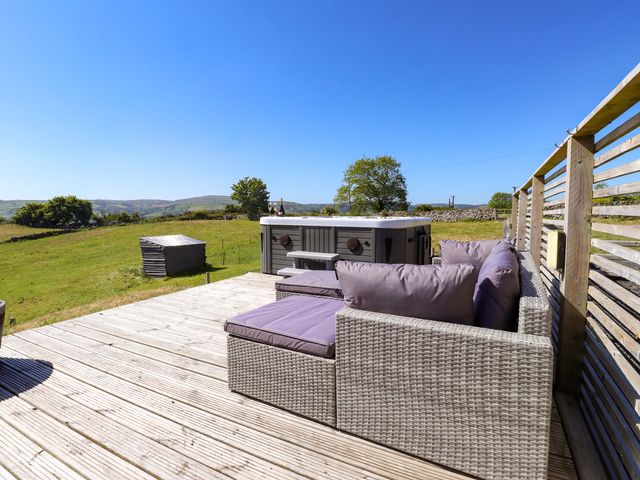 An outdoor wooden deck with a wicker sofa and a hot tub overlooking a grassy field at The Wren in Llysfaen
