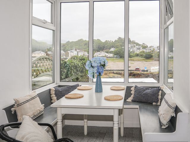 A dining nook with a white table and wicker placemats surrounded by black bench seating and cushions with a vase of blue flowers at Ivy Cottage in Borth-y-Gest near Porthmadog