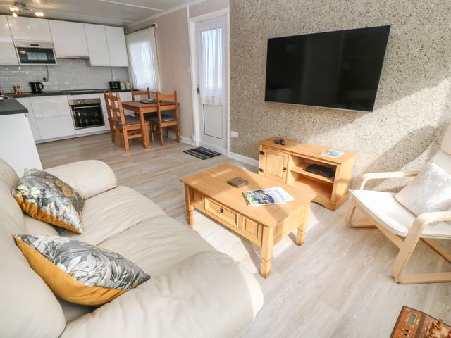 A living room with a beige sofa and cushions a wooden coffee table a wooden dining table with chairs a wall mounted television a wooden cabinet and a white armchair at Valhalla in Carbis Bay