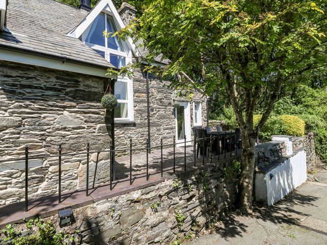 An outdoor patio area with a stone wall and railing outside a stone house with a tree and garden at Yr Hen Ysgol The Old School in Prenteg