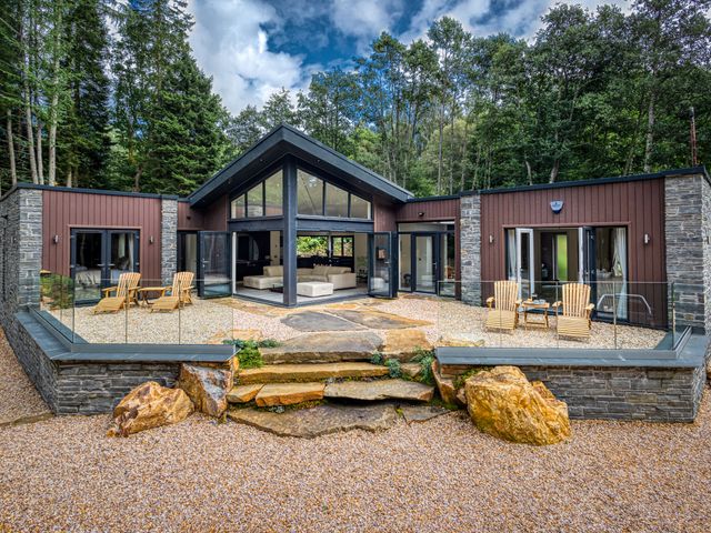 A modern house with glass doors and wooden chairs on a gravel patio surrounded by trees at Heilan Roo in Killiecrankie near Pitlochry and Blair Atholl