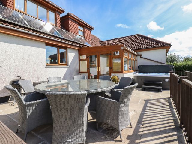 An outdoor patio with a round glass table and six wicker chairs next to a white house with solar panels and a hot tub at Meadowside in Goonhavern