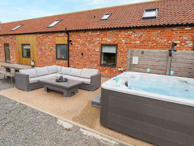 An outdoor patio area with a grey cushioned corner sofa and glass coffee table beside a hot tub and a red brick building at Pheasant's Roost in Lelley near Burton Pidsea