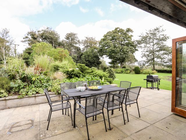 An outdoor patio with a black metal table and six chairs next to a garden and a lawn with trees and a barbecue grill at Clynnog House in Dwyran near Newborough