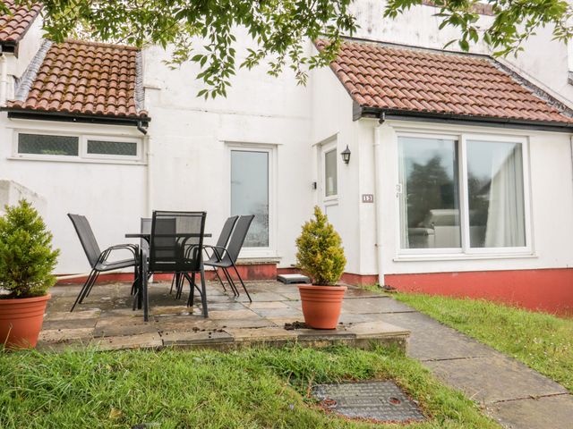 An outdoor patio with a table and six chairs in front of a white building with tiled roof and sliding glass door at 13 Manorcombe Bungalows in Honicombe Holiday Village near Gunnislake