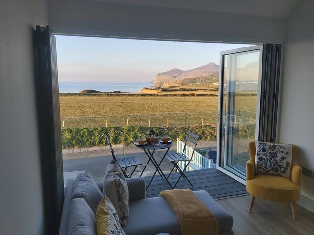 A living room with a gray sofa and yellow armchair opening to a balcony with table set and mountain view at Porth Penwaig in Nefyn
