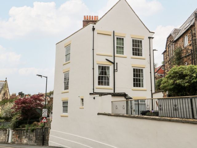 A white three-story house with multiple windows and a brick chimney next to a stone wall and greenery at Trillo House in Whitby