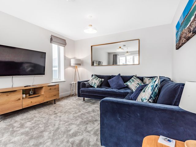 A living room with a blue sofa set a wooden TV stand a TV a floor lamp and a large mirror at Chi Gwynn in Crantock