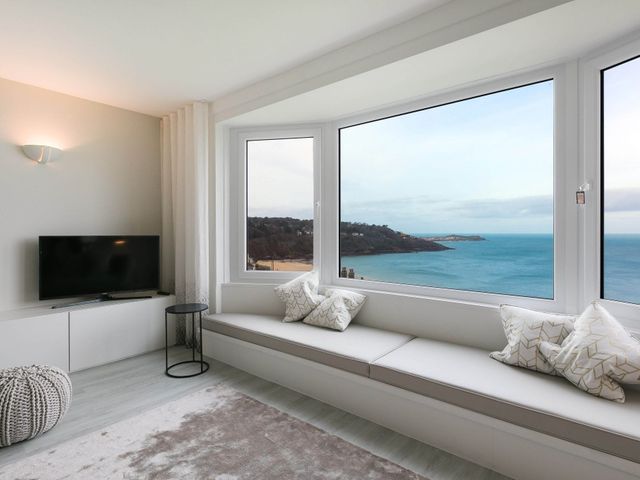 A living room with a wall mounted tv a round side table a knitted pouf window seat with cushions and sea view at Golden Heights in Carbis Bay