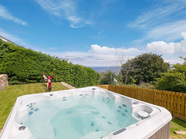 An outdoor hot tub on a lawn with green bushes and a wooden fence overlooking the sea at Bayside in Carbis Bay