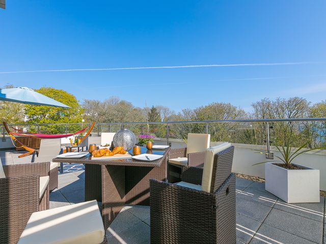 An outdoor patio with wicker chairs a table with croissants and cups a hammock and umbrella with trees and blue sky in the background at Sea Glass in Carbis Bay