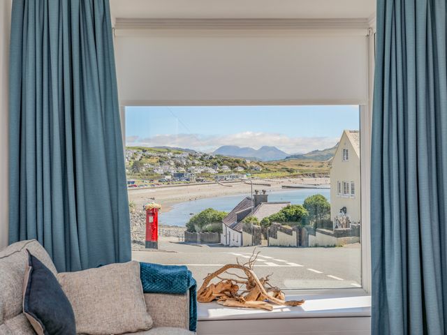 A window view of a coastal town with a red pillar box and houses from a living room with blue curtains and a beige sofa at Gwynle - Criccieth in Criccieth