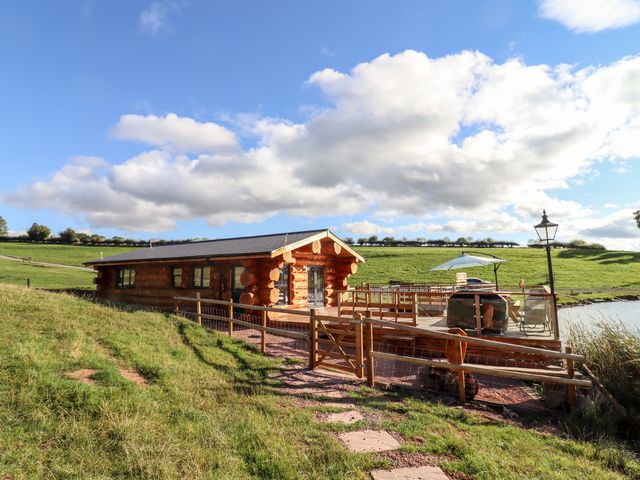 A log cabin with a wooden deck and outdoor furniture next to a lake in a grassy field near Clifton Upon Teme