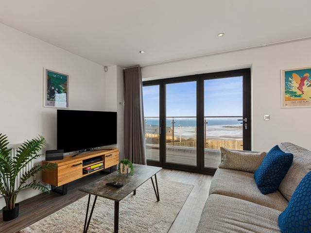 A living room with a sofa and cushions a coffee table a TV on a wooden stand and large glass doors showing a sea view at One Sand Banks in Broad Haven
