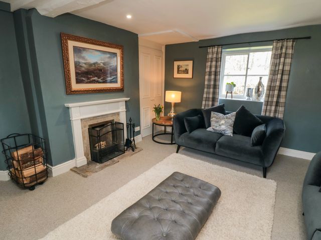 A living room with a fireplace a black sofa a tufted ottoman a table lamp and a window with plaid curtains at Black Horse Cottage in Swainby